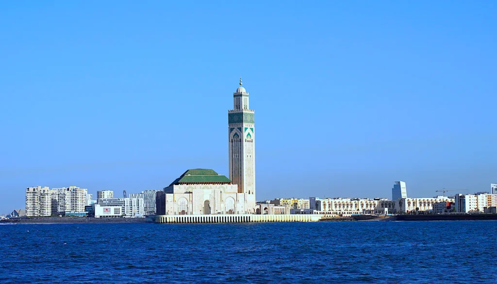 A scenic view of the Hassan II Mosque in Casablanca, Morocco, with its towering minaret, surrounded by modern city buildings and the Atlantic Ocean in the foreground