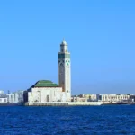 A scenic view of the Hassan II Mosque in Casablanca, Morocco, with its towering minaret, surrounded by modern city buildings and the Atlantic Ocean in the foreground