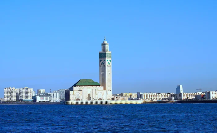 A scenic view of the Hassan II Mosque in Casablanca, Morocco, with its towering minaret, surrounded by modern city buildings and the Atlantic Ocean in the foreground