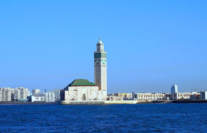 A scenic view of the Hassan II Mosque in Casablanca, Morocco, with its towering minaret, surrounded by modern city buildings and the Atlantic Ocean in the foreground