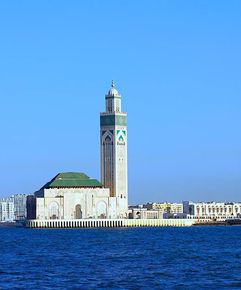 A scenic view of the Hassan II Mosque in Casablanca, Morocco, with its towering minaret, surrounded by modern city buildings and the Atlantic Ocean in the foreground