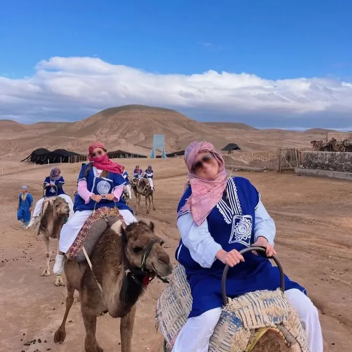 Tourists wearing traditional Moroccan clothing ride camels through the scenic Agafay Desert, with rolling hills and Berber tents in the background.