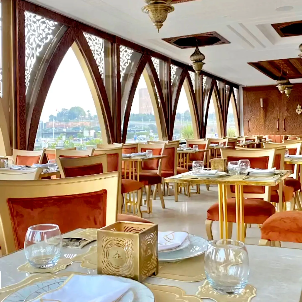 A beautifully decorated riverside restaurant with Moroccan-style arches, golden accents, and stunning views of the Hassan Tower in Rabat.