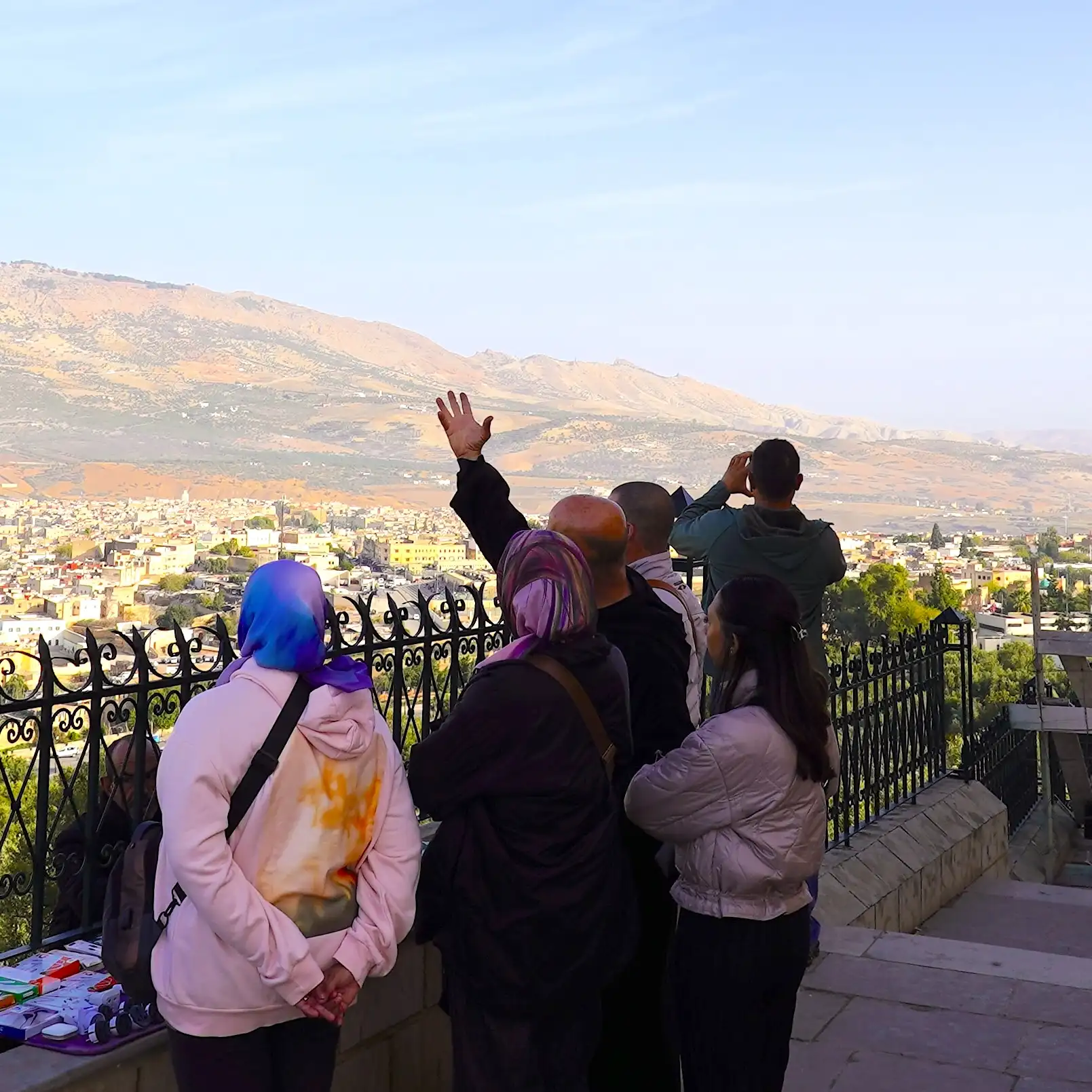 A guide explaining the historical significance of Fes to a group of tourists at a scenic overlook with a panoramic view of the city and hills