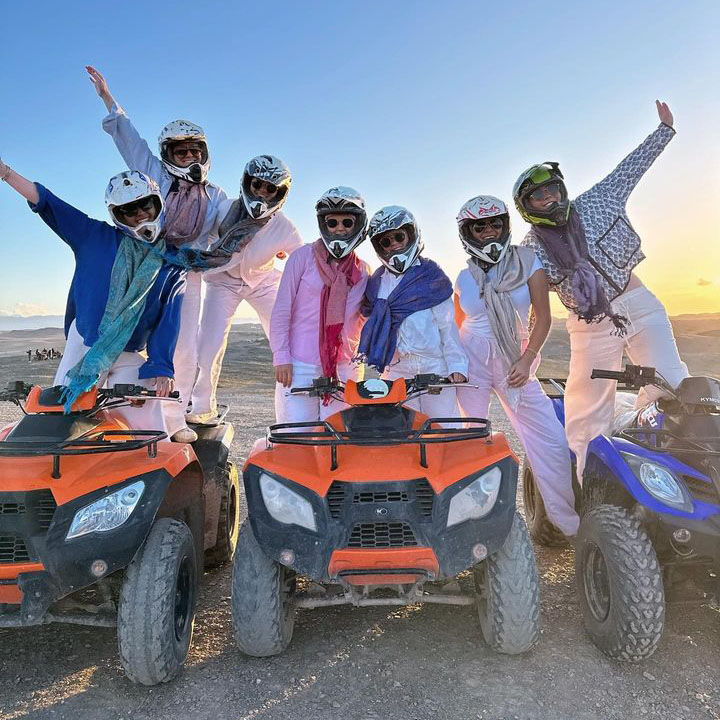 A group of people on quad bikes in the Agafay Desert, raising their hands in celebration under a bright blue sky with desert terrain in the background.