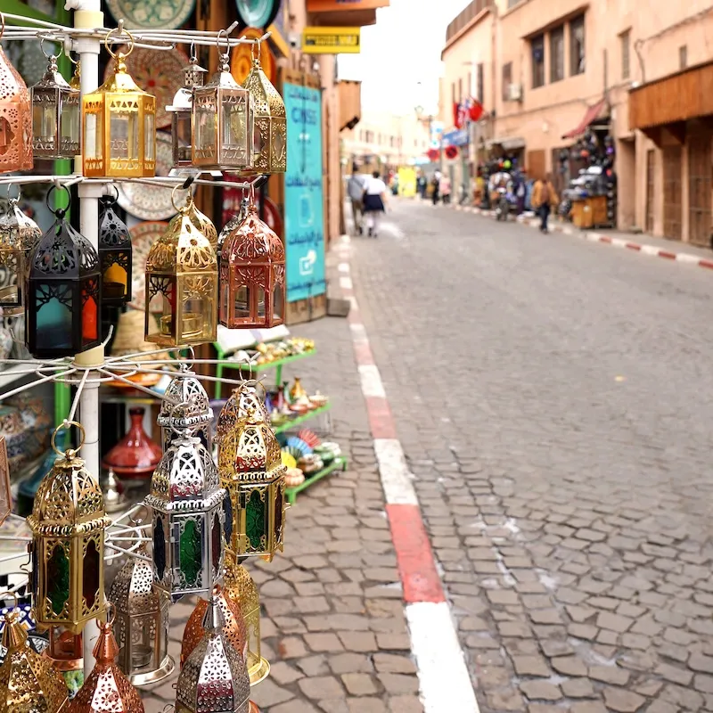 Traditional lanterns on display in the bustling streets of Marrakech’s Medina, with shops and pedestrians in the background.