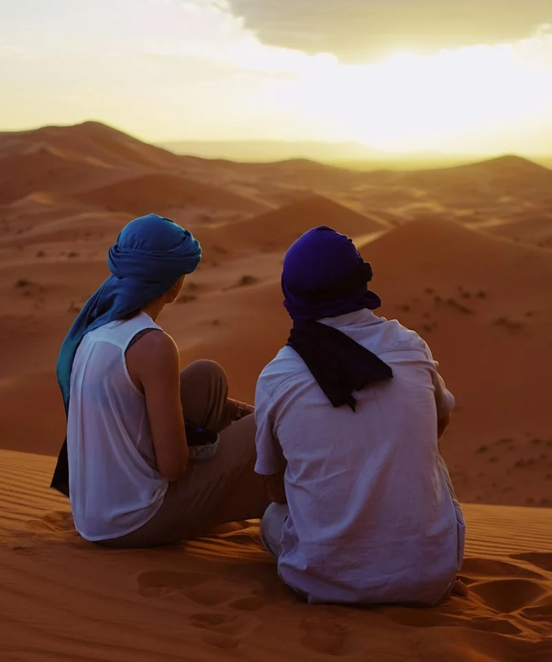 A couple sitting on golden sand dunes in Morocco, watching the sunset as the sky glows in warm hues, creating a romantic and peaceful atmosphere.