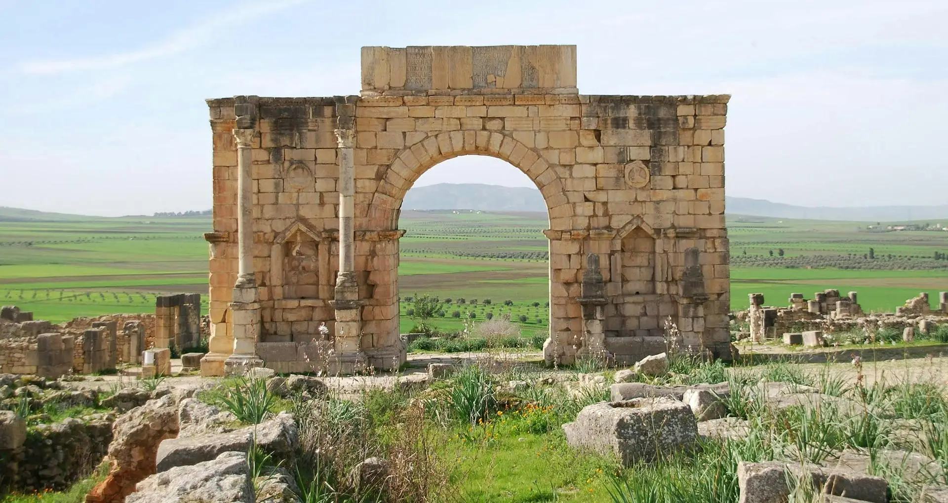 Ancient entrance arch at the Volubilis ruins near Meknes, Morocco, overlooking an open field with remnants of Roman architecture